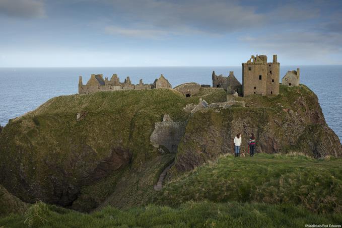Two women walking along a grassy clifftop, overlooking Dunnottar Castle, Aberdeenshire, situated on a rocky headland on the north-east coast of Scotland.