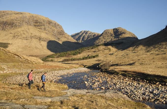 Two men hiking along a river in Glencoe, Scottish Highlands, mountains in the distance.