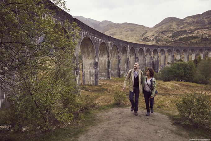 Glenfinnan Railway Viaduct, Highlands, Scotland. A mature couple walking on a footpath near the historic railway viaduct in an open landscape.