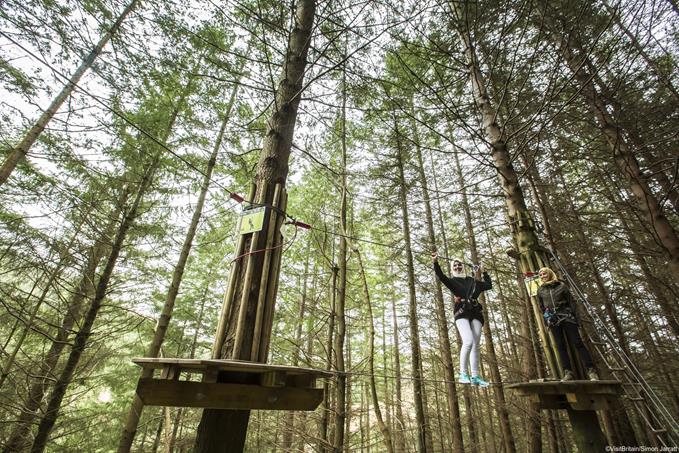 The Go Ape adventure park in Glentress Forest in the Tweed Valley. View from the ground of two young Asian women in headscarves on the forest treetop walkway, one walking on the wire between the platforms.