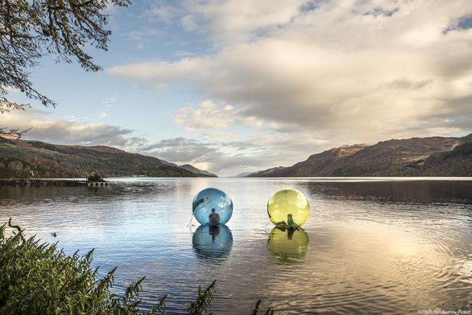 Two people zorbing, sitting in large plastic clear globes floating on the surface of Loch Ness. Flat calm water.